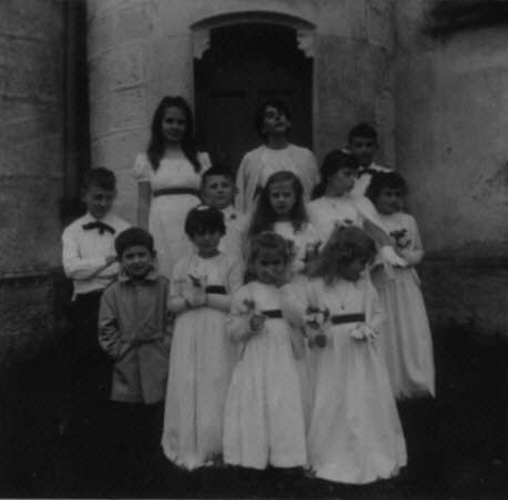 Photographie : un groupe de premiers communiants pose devant la tour d'un château, les plus petits devant les plus grands. Ils tous en aube blanche, les filles avec un bouquet de fleurs. Dans le groupe, sur le côté, un petit garçon se tient les mains dans les poches de son imperméable mastic.